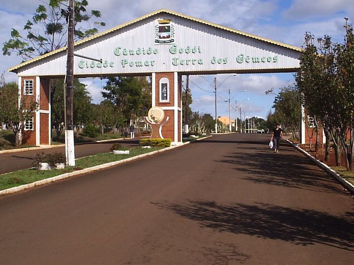 A white entrance arch over a road with green lettering reading Cândido Godói, Cidade Pomar, Terra dos Gêmeos, lined with trees under a blue sky