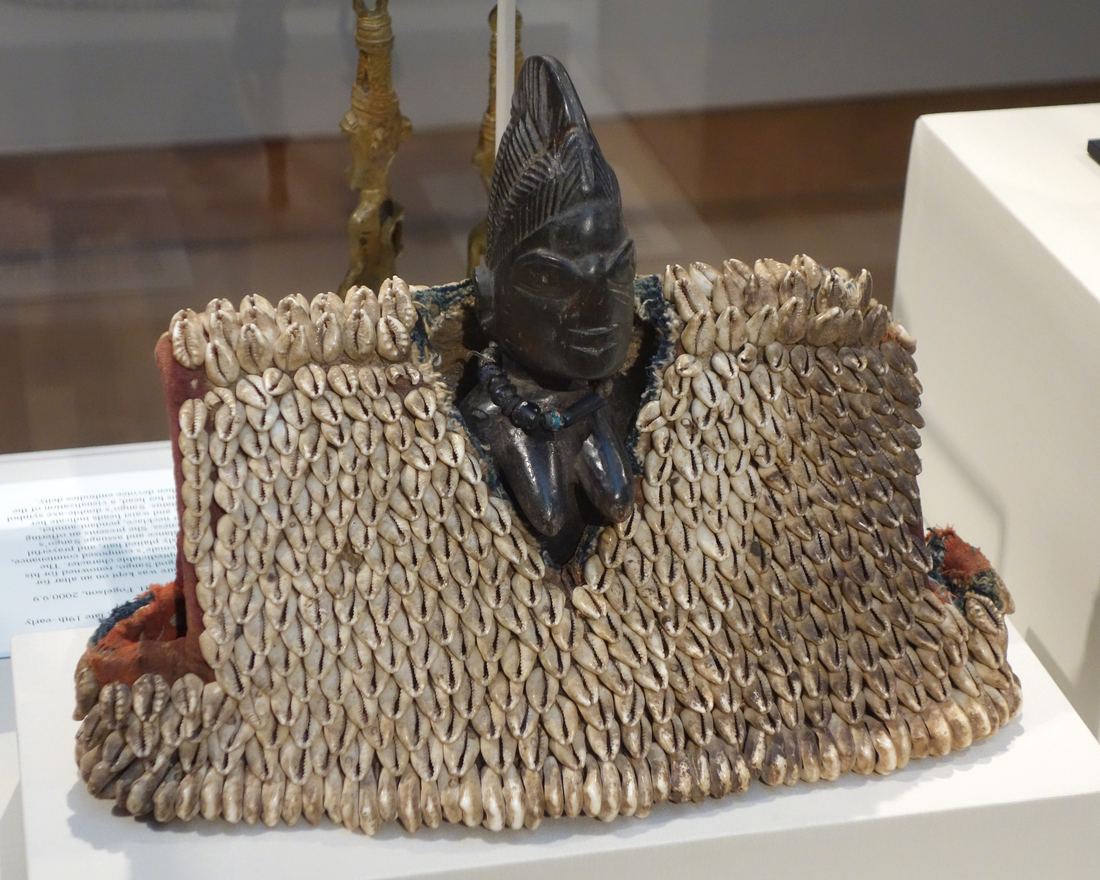 A small carved wooden Yoruba twin figure wrapped in a garment made of hundreds of cowrie shells, displayed in a museum case