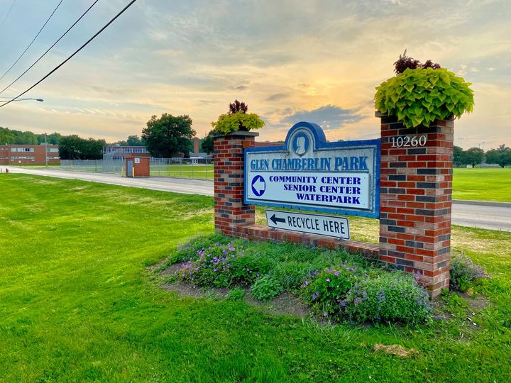 A brick and blue sign for Glen Chamberlin Park in Twinsburg, Ohio, with flower planters on top, set on a green lawn at sunset